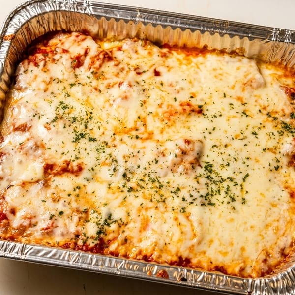 A large aluminum tray filled with baked lasagna, topped with melted cheese and herbs.