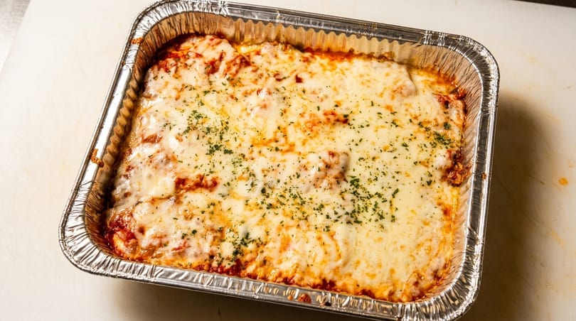 A large aluminum tray filled with baked lasagna, topped with melted cheese and herbs.