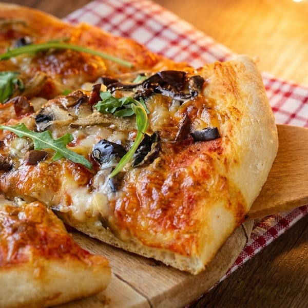 A slice of mushroom and olive pizza with arugula being lifted from a wooden board.
