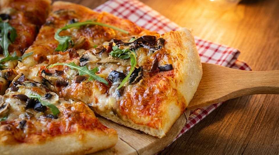 A slice of mushroom and olive pizza with arugula being lifted from a wooden board.